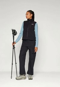 Γυναίκα με εξωτερική ένδυση κρατώντας διπλωμένα ραβδιά πεζοπορίας, φορώντας μαύρο γιλέκο, μπλε μακρυμάνικο μπλουζάκι, μαύρα παντελόνια cargo και γκρι αθλητικά παπούτσια.