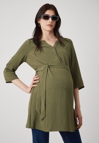 Vihreä oliivinvärinen äitiysmekko, jossa on V-pääntie, kolmannes hihoista, napitusyksityiskohtia ja sidottu vyötärö, sekä käytännöllinen tekstuuri.