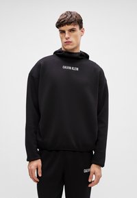 Čierna mikina s kapucňou a voľným strihom, na prednej strane s bielym nápisom "CALVIN KLEIN". Vyrobená z mäkkého materiálu bez vzorov.