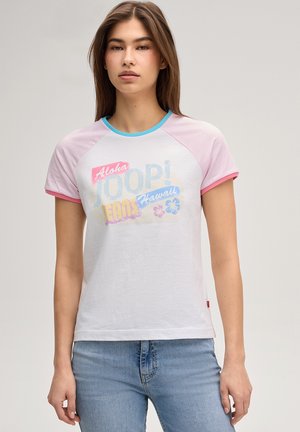 Młoda kobieta w białej i różowej koszulce z raglanowymi rękawami z napisem „Aloha JOOP! Jeans Hawaii” oraz jasnoniebieskich dżinsach, stojąca neutralnie.