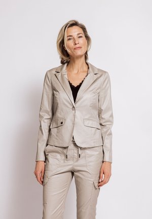 Woman with short blonde hair wearing a beige jacket and matching pants over a black top, standing against a plain light background.