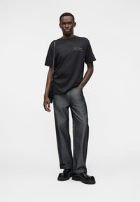 Μαύρο βαμβακερό T-shirt με μινιμαλιστικό σχέδιο και κεντημένο κείμενο, σε συνδυασμό με γκρι φαρδιά τζιν παντελόνια και χοντρά μαύρα παπούτσια.