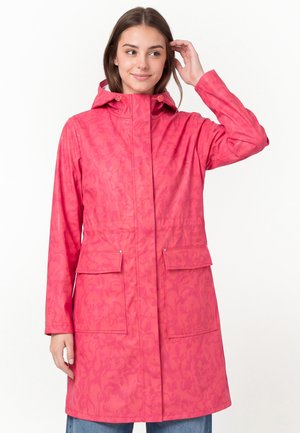 Ung kvinde iført en lang pink regnjakke med hætte, der holder håret tilbage med den ene hånd og står mod en hvid baggrund.