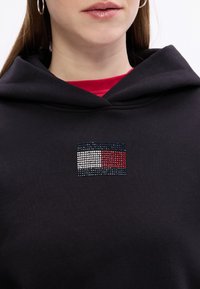 Czarna bluza z kapturem z logo osadzonym w kryształkach w prostokątnym wzorze, zawierającym niebieskie, białe i czerwone sekcje.
