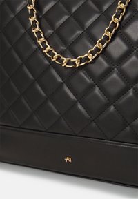 Bolsa de mano acolchada negra de cuero suave, con una correa de cadena dorada y un pequeño acento de logo dorado en la base. Superficie texturizada con patrón de diamantes.