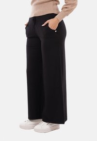 Persona che indossa pantaloni larghi neri con tasche, un maglione beige e sneaker bianche, in piedi con le mani nelle tasche.