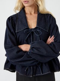 Mulher vestindo uma blusa de denim azul-escuro com um grande colarinho, fecho em laço e mangas longas bufantes com punhos elásticos, com os braços cruzados.