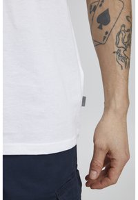 Hvid bomulds T-shirt med lige pasform, der har en synlig søm, sidesømsetiket og kontrasterende mørkeblå shorts, der viser arm tatoveringer.