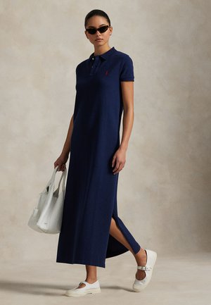 Μπλε σκούρο φόρεμα πόλο με κοντά μανίκια, πλαϊνή σχισμή και κόκκινο λογότυπο ως διακόσμηση. Συνδυασμένο με λευκά παπούτσια και λευκή τσάντα, με ουδέτερο φόντο.