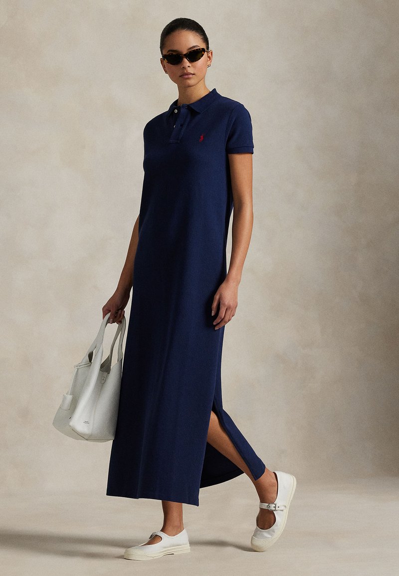 Mörkblå poloklänning med korta ärmar, en sidospalt och en röd logotypdetalj. Kombinerad med vita skor och en vit väska, mot en neutral bakgrund.