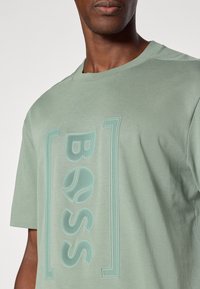 Tricou din bumbac verde deschis cu un design texturat al logo-ului într-o nuanță mai deschisă, cu guler rotund și mâneci scurte.