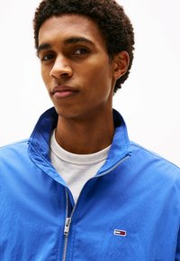 Giacca blu con zip e una texture morbida, caratterizzata da un colletto alto e un logo ricamato. Indossata sopra una camicia bianca.