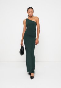Φόρεμα midi με ένα ώμο, εφαρμοστό, σε σκούρο πράσινο χρώμα, φτιαγμένο από ελαστικό ύφασμα. Το φόρεμα έχει κομψή σιλουέτα, συνδυασμένο με μαύρα αξεσουάρ.