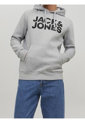 Osoba ubrana w jasnoszarą bluzę z kapturem z dużym czarnym napisem "JACK & JONES" oraz niebieskie dżinsy, ręce w przedniej kieszeni bluzy.