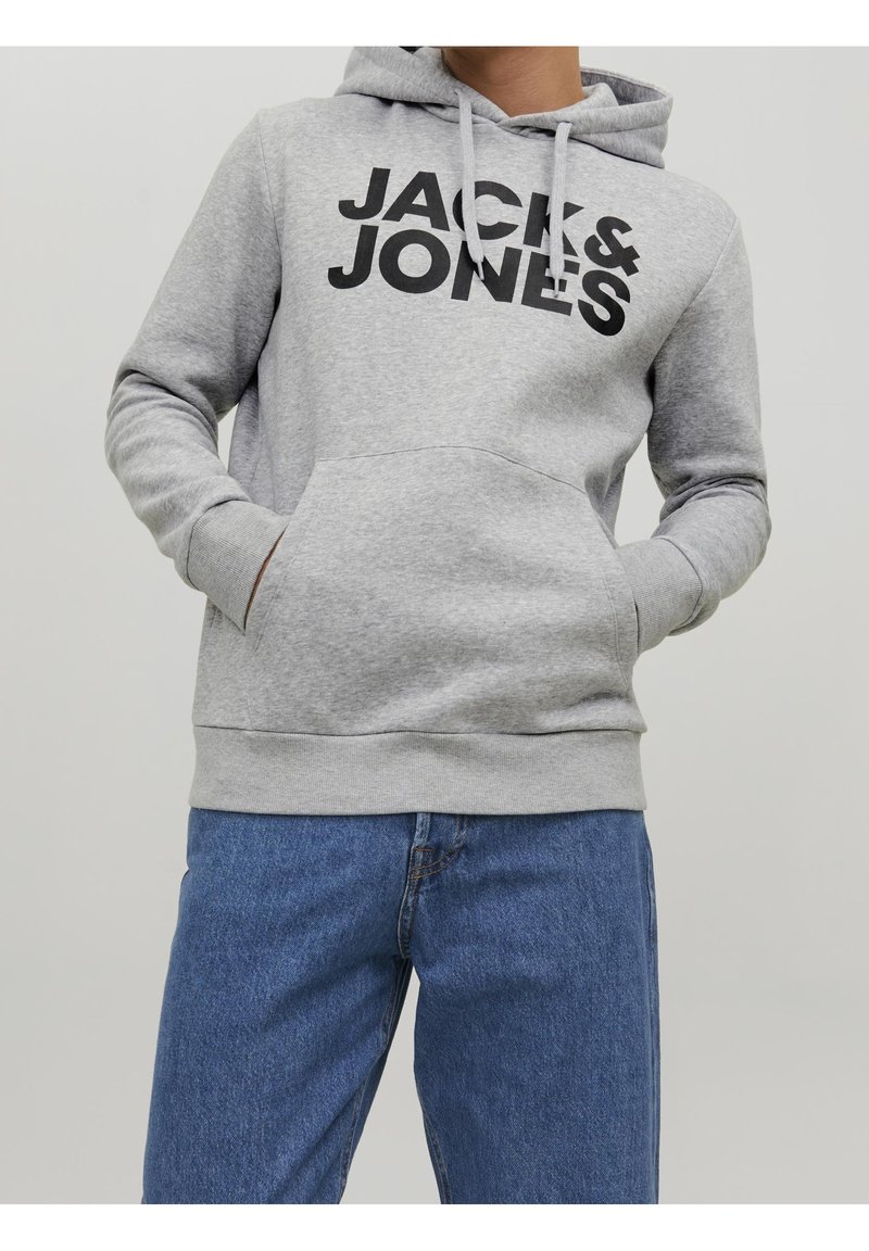 Personne portant un sweat à capuche gris clair avec un grand texte noir "JACK & JONES" et un jean bleu, mains dans la poche avant du sweat.