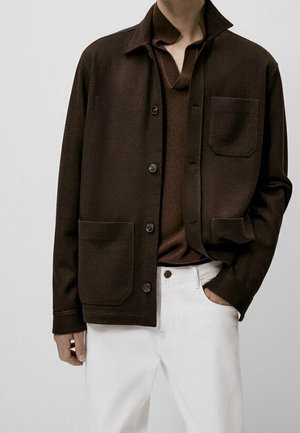 Mężczyzna w ciemnobrązowej kurtce na guziki z dużymi kieszeniami, nałożonej na pasującą brązową koszulę i białe spodnie, z ręką w kieszeni.