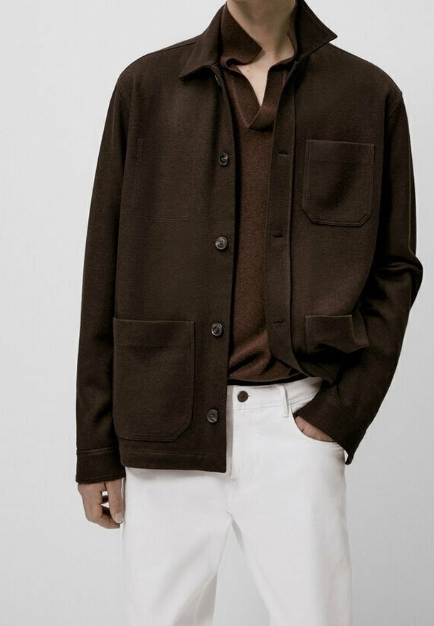 Homme portant une veste boutonnée marron foncé avec de grandes poches, sur une chemise marron assortie et un pantalon blanc, main dans la poche.