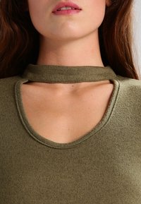 Olijfgroene top met een ronde hals en een open nekdetail. De stof is textuur en zacht, met zichtbare stiksels langs de randen.