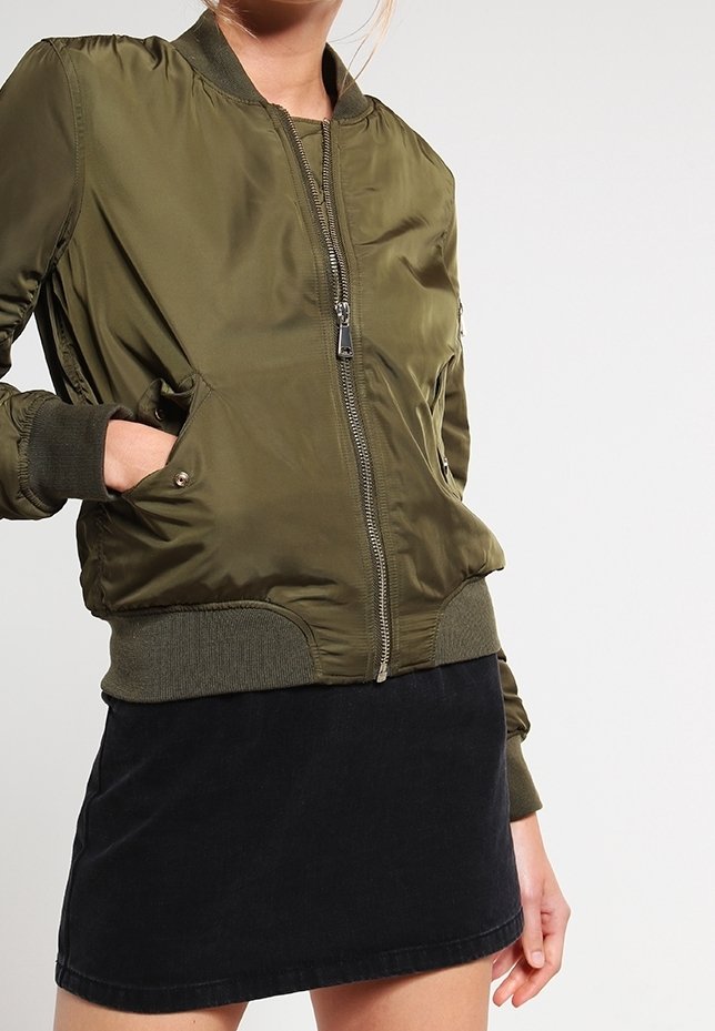 Personne portant une veste bomber vert olive avec fermeture éclair et poignets et ourlet côtelés, associée à une jupe noire sur un fond uni.