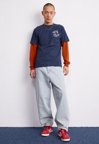 T-shirt azul-marinho sobre uma camisola laranja de manga longa, combinado com jeans baggy de tom azul claro e ténis vermelhos com detalhes brancos.