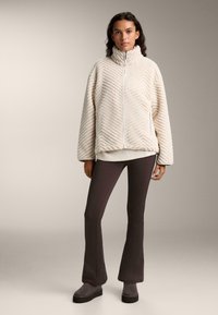 Cremefarbener Fleece-Pullover mit einem strukturierten Zickzack-Muster, hohem Kragen und Frontreißverschluss. Kombiniert mit dunklen Leggings und grauen Stiefeln.