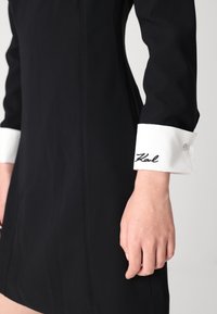 Črna obleka z dolgimi rokavi, belimi kontrastnimi manšetami s gumbom in izvezenim poudarkom "Karl" na manšeti, gladka tekstura tkanine.