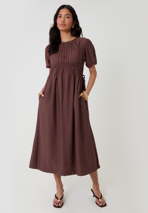 Frau trägt ein braunes Midikleid mit Puffärmeln und gerafftem Oberteil, steht mit den Händen in den Taschen und trägt braune Sandalen.
