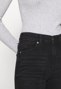 Lähiaufnahme von einer Person, die ein geripptes, hellgraues Langarmshirt trägt, das in hochgeschnittene schwarze Jeans mit vorderen Taschen und Knopfdetails gesteckt ist.