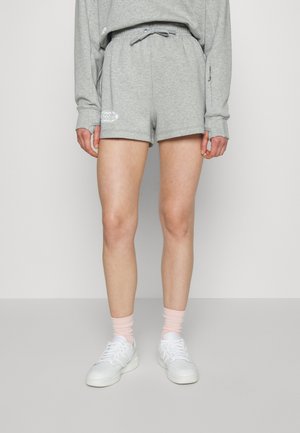 Personne portant un short gris clair à cordon, un haut à manches longues gris clair, des chaussettes rose pâle et des baskets blanches, debout devant un fond uni.