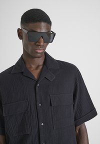 Zwart kortom shirt met een gestructureerd patroon, twee voorzakken en een knoopsluiting, gecombineerd met hoekige zwarte zonnebril.