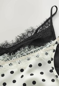 Deux camisoles ornées de dentelle, une noire et une blanche avec des pois noirs, posées à plat sur un fond clair.