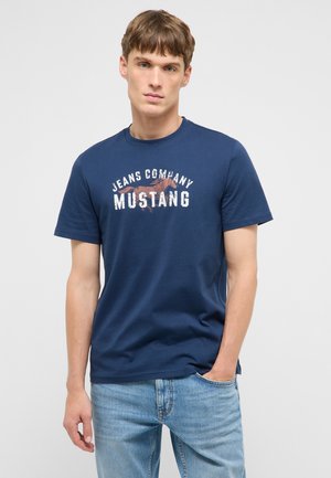 Junger Mann mit hellbraunen Haaren, der ein marineblaues "Jeans Company Mustang" T-Shirt und hellblaue Jeans trägt, steht mit einer Hand in der Tasche.