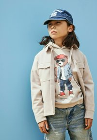 Enfant portant une casquette bleue, une veste beige et un sweat-shirt avec un ours portant des lunettes de soleil, debout devant un fond bleu.