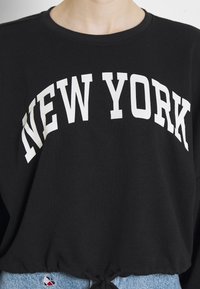 Czarna bluza z kapturem z napisem "NEW YORK" na piersi wykonanym dużymi białymi literami. Posiada sznurki do regulacji dolnej części.