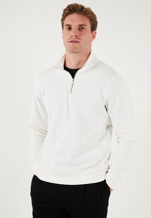 White zip-up sweatshirt with a high collar, soft fabric, long sleeves, and a relaxed fit, paired with black pants.