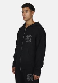 Młody mężczyzna ubrany w czarną bluzę z zamkiem i dopasowane spodnie z szarym logo, stojący na tle czystego białego tła.