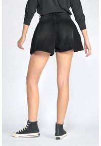 Osoba oblečená v černých džínových šortkách s vysokým pasem, černém topu s dlouhým rukávem a černých vysokých teniskách, zobrazena zezadu na bílém pozadí.