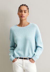 Helles blaues strukturiertes Sweatshirt mit weit ausgeschnittenem Hals, langen Ärmeln und geripptem Saum, kombiniert mit weißen Hosen und einem schwarzen Gürtel.