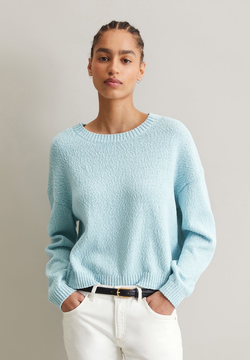 Helles blaues strukturiertes Sweatshirt mit weit ausgeschnittenem Hals, langen Ärmeln und geripptem Saum, kombiniert mit weißen Hosen und einem schwarzen Gürtel.