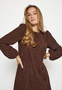 Hnedé štruktúrované šaty s dlhými puff rukávmi a okrúhlym výstrihom. Obsahujú bočné vrecká a voľný strih. Mäkka tkanina s jemným vzorom.