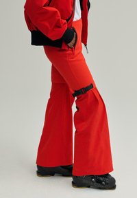 Pantaloni evazați roșii cu talie înaltă, cu un detaliu negru reglabil pe coapsă. Materialul pare neted și structurat.