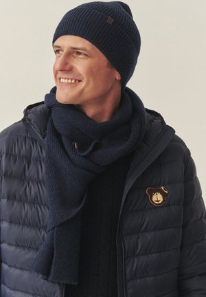 Căciulă și fular tricotate cu model în relief, de culoare bleumarin, asortate cu o jachetă de tip puffer închisă la culoare, cu o emblema cu urs. Materiale călduroase și modele texturate sunt vizibile.