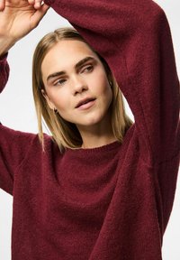 Burgundiin neulepaita, jossa on leveä pääntie, pitkät hihat ja tekstuuripintainen viimeistely. Pehmeä, mukava kangas laskeutuu löyhästi kehon ympärille.