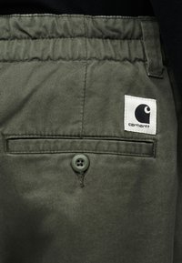 Πράσινο παντελόνι από ύφασμα με κουμπωτή πίσω τσέπη και λευκή ετικέτα λογότυπου Carhartt κοντά στη μέση.