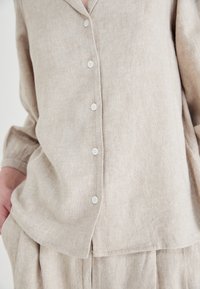 Camicia di lino beige con scollatura a V, maniche lunghe e cinque bottoni bianchi lungo la parte anteriore. Il tessuto ha un aspetto morbido e testurizzato.