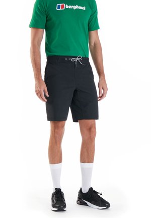 Zwarte zwembaden met een elastische tailleband en een trekkoord, gecombineerd met een groene T-shirt met een wit logo, zwarte schoenen en witte sokken.