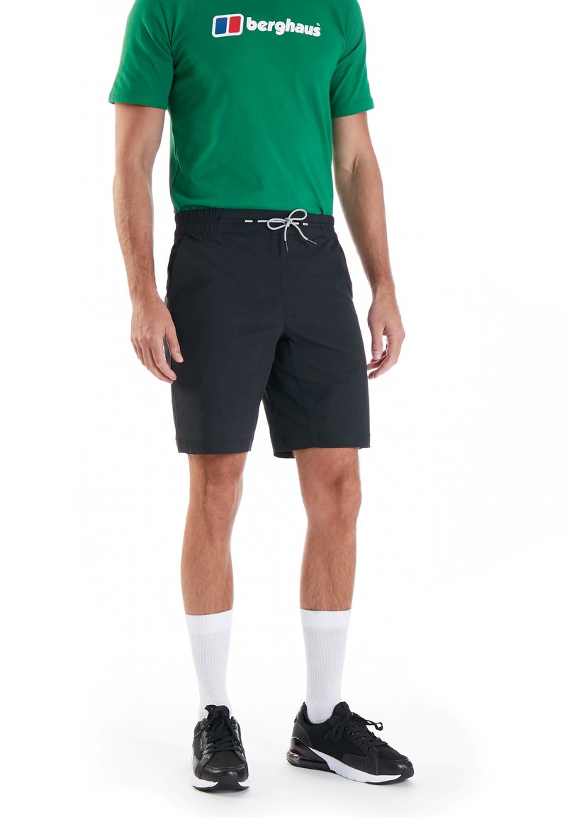 Schwarze Badehose mit einem elastischen Bund und Kordelzug, kombiniert mit einem grünen T-Shirt mit einem weißen Logo, schwarzen Schuhen und weißen Socken.