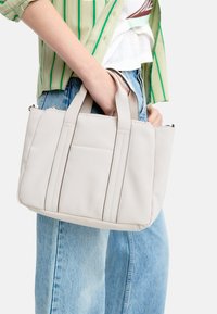 Jasnoszara torba na ramię z podwójnymi uchwytami, zamknięciem na suwak, przeszyciami oraz boczną kieszenią. Materiał wydaje się gładki i trwały.