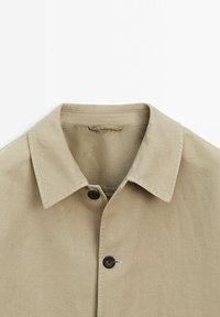 Camicia in cotone beige con colletto a punta, dotata di un bottone nero e un dettaglio ad anello vicino al bottone per la chiusura. Superficie del tessuto texture.
