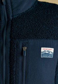 Tummansininen fleece-takki, jossa on teksturoitu kangas, edessä vetoketju, vetoketjullinen rintatasku ja Joules Outdoor Apparel -merkki, jossa on vuoria.